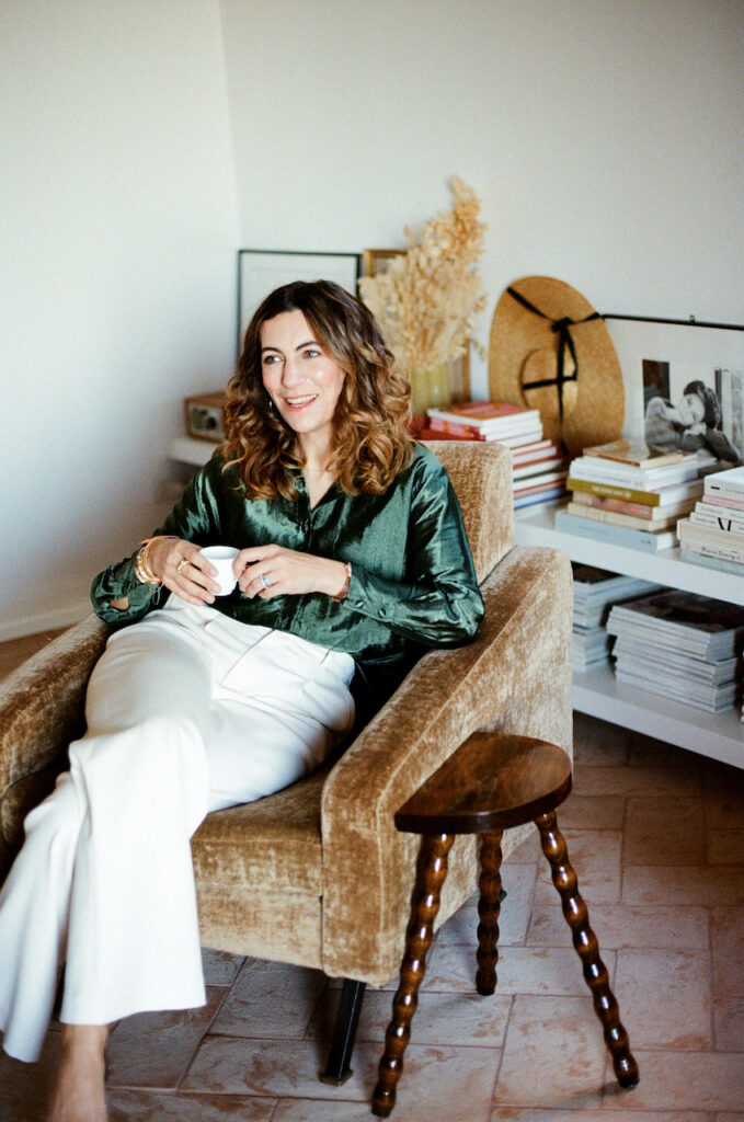 Alice Chéron assise dans un fauteuil en velours moutarde, intérieur chaleureux avec tomettes et étagères de livres d'art.