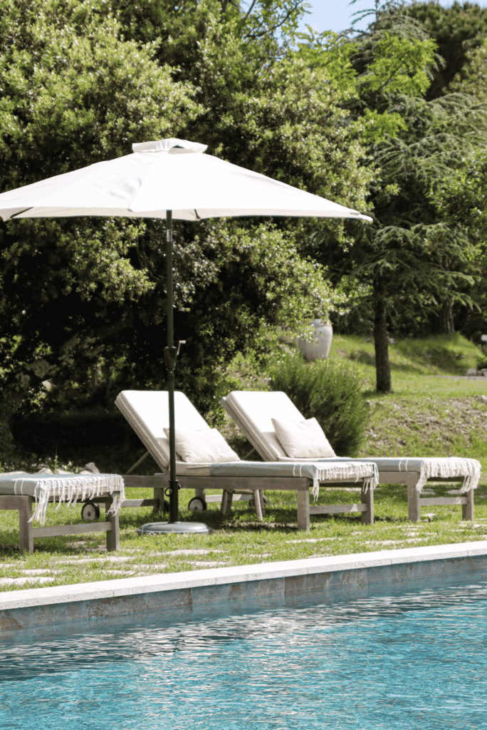 Piscine aux eaux turquoise bordée de transats en bois avec coussins blancs et parasol, entourée de verdure.