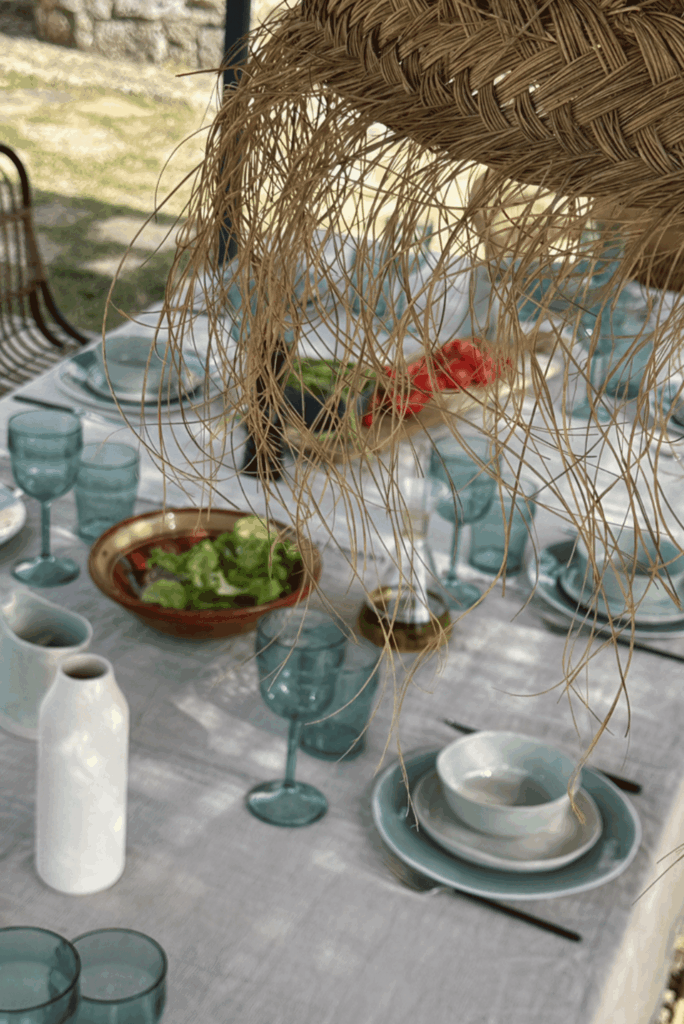 Table dressée avec nappe en lin, verres bleus et vaisselle artisanale sous un luminaire en paille tressée.