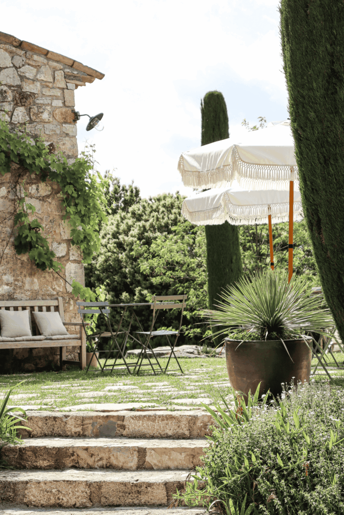 Terrasse ombragée au Domaine de la Xavolière avec parasols à franges blancs, mobilier en fer et végétation méditerranéenne.