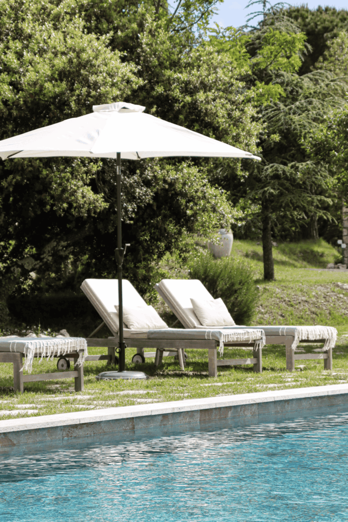 Piscine aux eaux turquoise bordée de transats en bois avec coussins blancs et parasol, entourée de verdure.