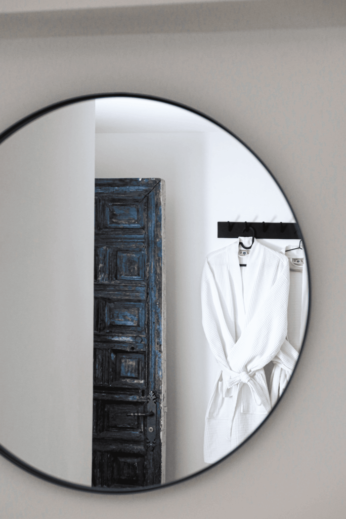 Reflet dans un miroir rond noir d'une porte ancienne en bois bleu délavé et d'un peignoir blanc suspendu.